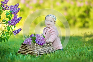 Smiling baby girl 1-2 year old wearing flower wreath, holding bouquet of lilac outdoors. Looking at camera. Summer spring time.