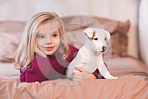Smiling baby girl 3-4 year old lying in bed with puppy dog of jack russel terrier close up. Friendship.
