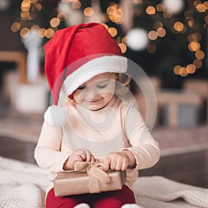 Smiling baby girl 3-4 year old open Christmas present box  wearing red santa claus hat and pajamas over glowing lights close up.