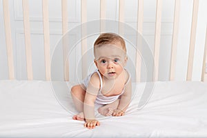 Smiling baby girl 8 months old sitting in a crib in a children`s room in white clothes and looking at the camera, baby`s morning