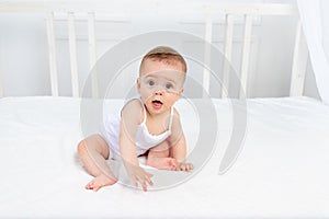 Smiling baby girl 8 months old sitting in a crib in a children`s room in white clothes and looking at the camera, baby`s morning
