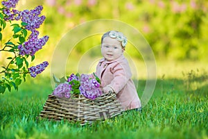 Smiling baby girl 1-2 year old wearing flower wreath, holding bouquet of lilac outdoors. Looking at camera. Summer spring time.