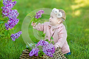 Smiling baby girl 1-2 year old wearing flower wreath, holding bouquet of lilac outdoors. Looking at camera. Summer spring time.