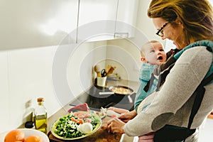 Smiling baby ported in baby carrier backpack looking at his mother while she cooks, concept of family conciliation