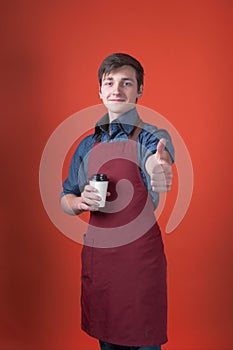 Smiling barista with dark hair in red apron holding coffee in paper cup, looking at camera and thumbing up