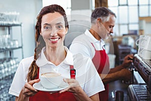 Smiling barista holding a cup of coffee with colleague behind