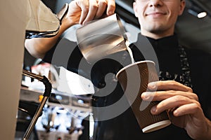 Smiling barista man pouring whipped milk from frothing pitcher in paper cup with coffee standing in front of