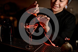 Smiling bartender girl sprinkling on a alcoholic drink in the cocktail glass with a orange zest juice