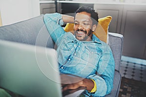 Smiling bearded African man spending free time in sofa and using digital tablet at modern home.Concept of young people