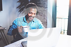 Smiling bearded African man using laptop home while drinking cup black coffee at the wooden table.Concept of young