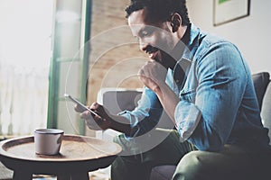 Smiling bearded African man using smartphone while sitting on sofa at home.Concept people working with mobile gadget