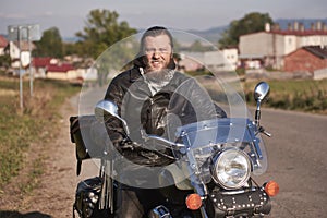 Smiling bearded biker in black leather jacket sitting on modern motorcycle on country roadside.