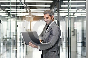 Smiling bearded business man focused on company datas while standing in office corridor and using laptop.