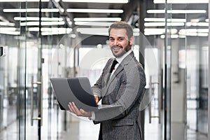 Smiling bearded business man focused on company datas while standing in office corridor and using laptop.