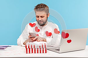 Smiling bearded businessman in white shirt in red heart shaped stickers sitting at desk with laptop and striped giftbox typing on