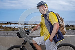 Smiling bearded cyclist senior man along the sea beach carrying a backpack, wearing helmet running with electro bicycle looking at