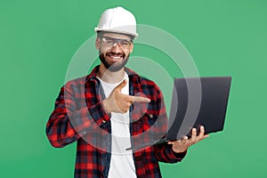 Smiling bearded engineer or constructor man in casual outfit pointing finger on computer over green background.
