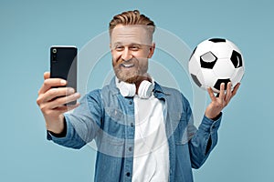 Smiling bearded hipster blonde young man take selfie on smartphone holding classic soccer ball over blue background.