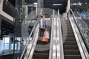 Smiling bearded man carrying shopping bags, while going down on escalator of shopping mall.