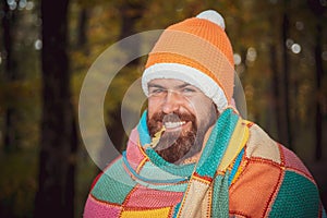 Smiling bearded man holds autumnal oak leaf in his teeth. Hipster guy wearing knitted orange hat and cozy warm plaid