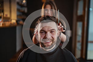 Smiling bearded Man portrait while undercut hairstyle hairdressing in Hair Salon by young hairdresser female. Modern low light