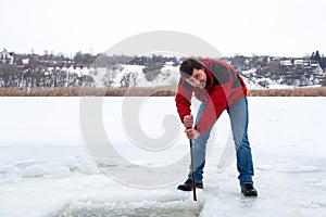 Smiling bearded man in a red jacket and jeans pierces the ice on the frozen river with a crowbar