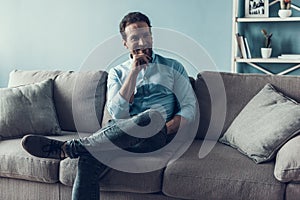 Smiling Bearded Man Sitting on Gray Sofa at Home.