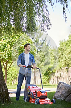 Smiling bearded man trimming lawn with modern mower, while working in garden at sunny day.