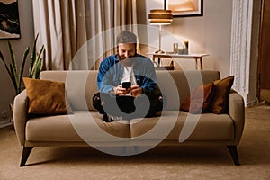 Smiling bearded man using mobile phone while sitting in lotus pose on sofa at home