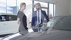 Smiling bearded salesman in business suit showing new auto to successful business woman at car dealership. Showroom