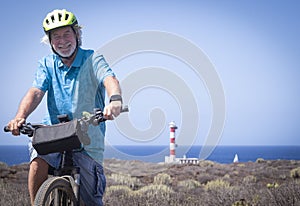 Smiling bearded senior man with yellow helmet enjoying riding bicycle close to a lighthouse - arid landscape and horizon over