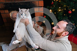 Smiling bearded young man holding in hands cute fluffy cat surrounded by family of wife and little son by Christmas tree