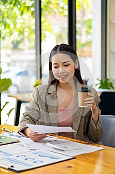 Smiling beautiful Asian businesswoman holding coffee cup and laptop working on graph papers at financial accounting concept office