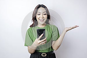 Smiling beautiful Asian woman pointing finger sideward to empty space while holding her phone isolated white background