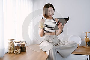 Smiling beautiful Asian woman sits and relaxes on top counter kitchen using tablet browsing unlimited wireless internet in kitchen