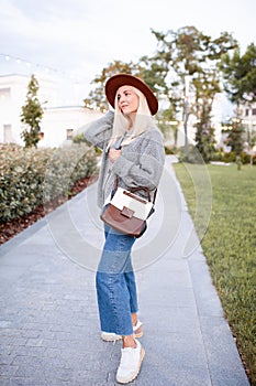 Smiling beautiful blonde teenage girl 17-18 year old wear stylish hat, leather bag and knit gray jumper walking on city street