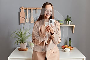 Smiling beautiful business woman with brown hair wearing beige jacket standing in home kitchen interior using mobile phone