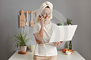 Smiling beautiful busy young woman posing in kitchen with towel on head doing beauty treatment procedures talking phone working