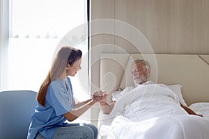 Smiling beautiful caring female doctor holding hand of male senior patient who lying in hospital bed. Nurse takes care elderly man