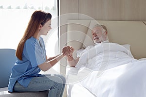 Smiling beautiful caring female doctor holding hand of male senior patient who lying in hospital bed. Nurse takes care elderly man