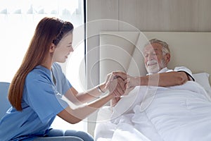 Smiling beautiful caring female doctor holding hand of male senior patient who lying in hospital bed. Nurse takes care elderly man