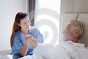 Smiling beautiful caring female doctor holding hand of male senior patient who lying in hospital bed. Nurse takes care elderly man