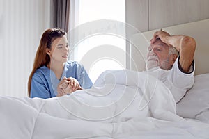 Smiling beautiful caring female doctor holding hand of male senior patient who lying in hospital bed. Nurse takes care elderly man