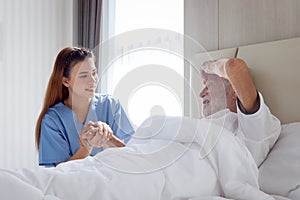 Smiling beautiful caring female doctor holding hand of male senior patient who lying in hospital bed. Nurse takes care elderly man