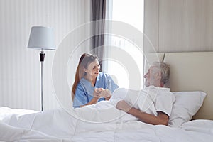 Smiling beautiful caring female doctor holding hand of male senior patient who lying in hospital bed. Nurse takes care elderly man