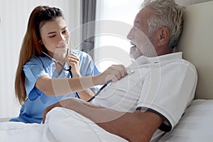 Smiling beautiful caring female doctor using stethoscope listen to senior patient heart who lying in hospital bed. Nurse takes