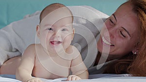 Smiling beautiful Caucasian mother playing with newborn baby boy on the bed in bedroom. Parent and cute infant childhood