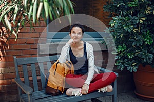 Smiling beautiful Caucasian young girl woman in white sweater and red jeans, sitting with yellow travel bag backpack on bench