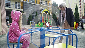 Smiling beautiful grandmother swinging merry-go-round with cute little granddaughter sitting on revolving platform. Wide