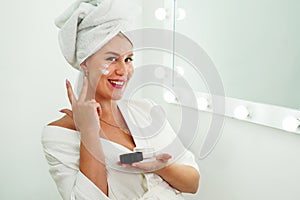 Smiling beautiful lady apply skincare cream on face look in bathroom mirror, happy young woman wrap towel on head put